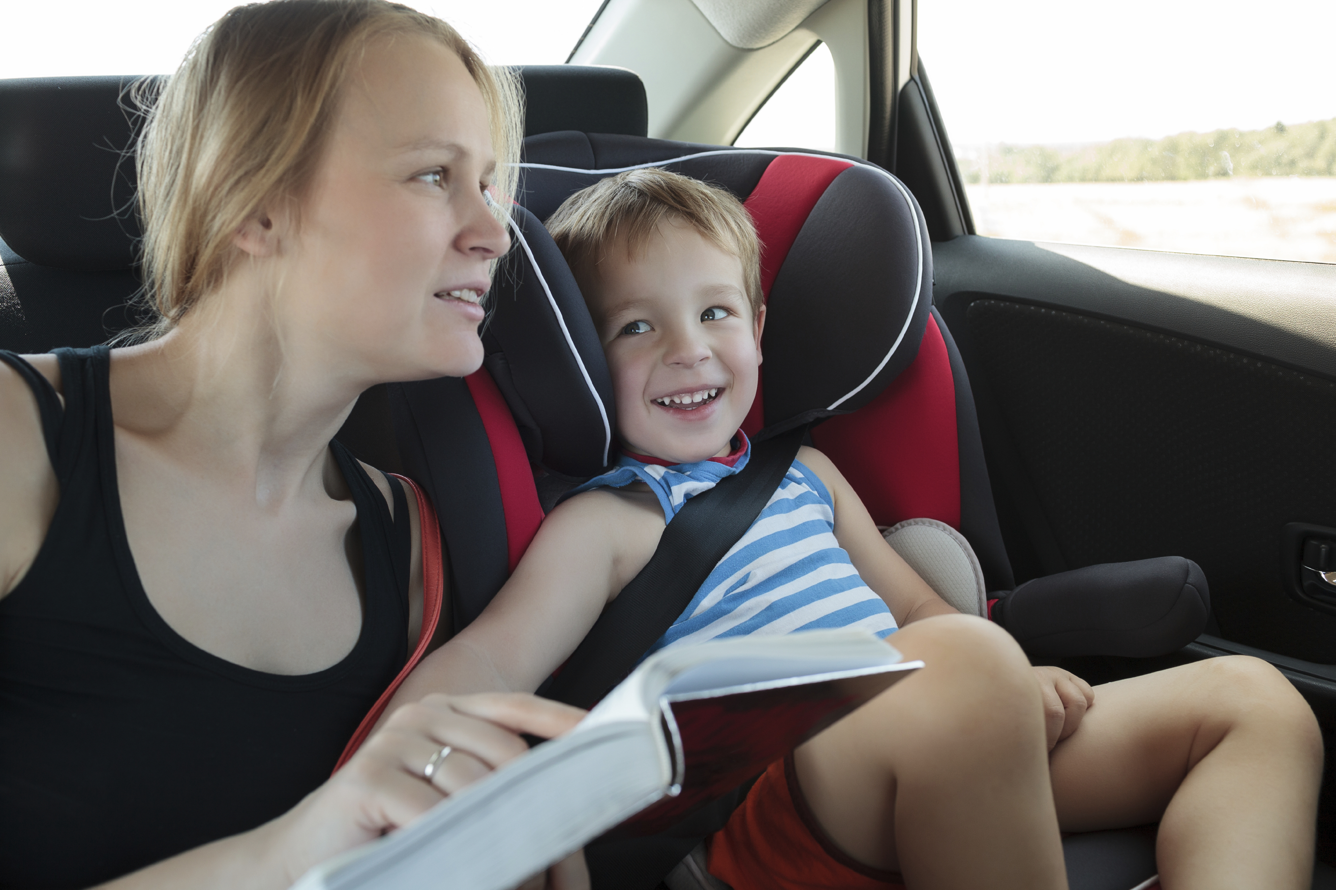 Mother and son traveling by car. Woman reading a book to the kid sitting in child safety seat